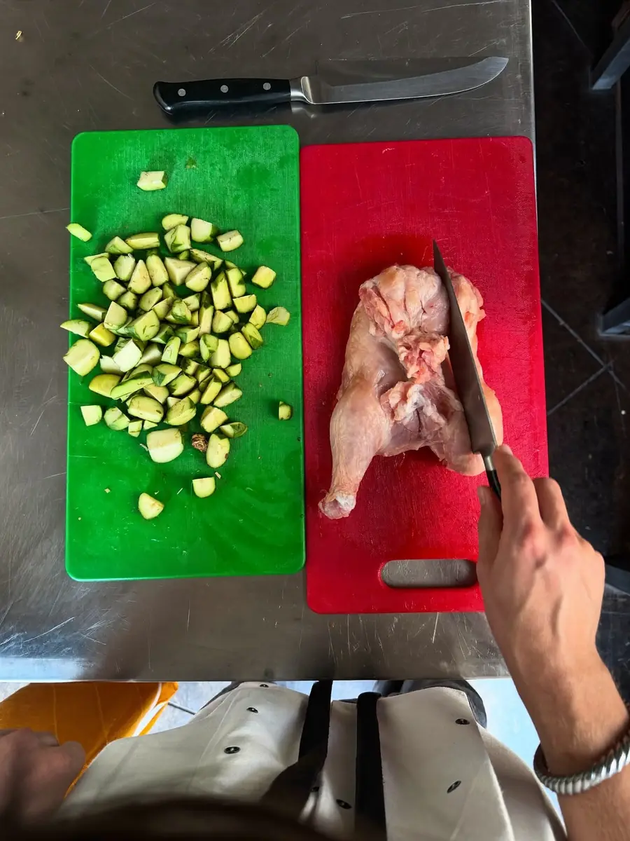 Overhead view of a red chopping board holding raw chicken beside a green board of chopped vegetables on a stainless steel bench, kept separate to prevent cross-contamination